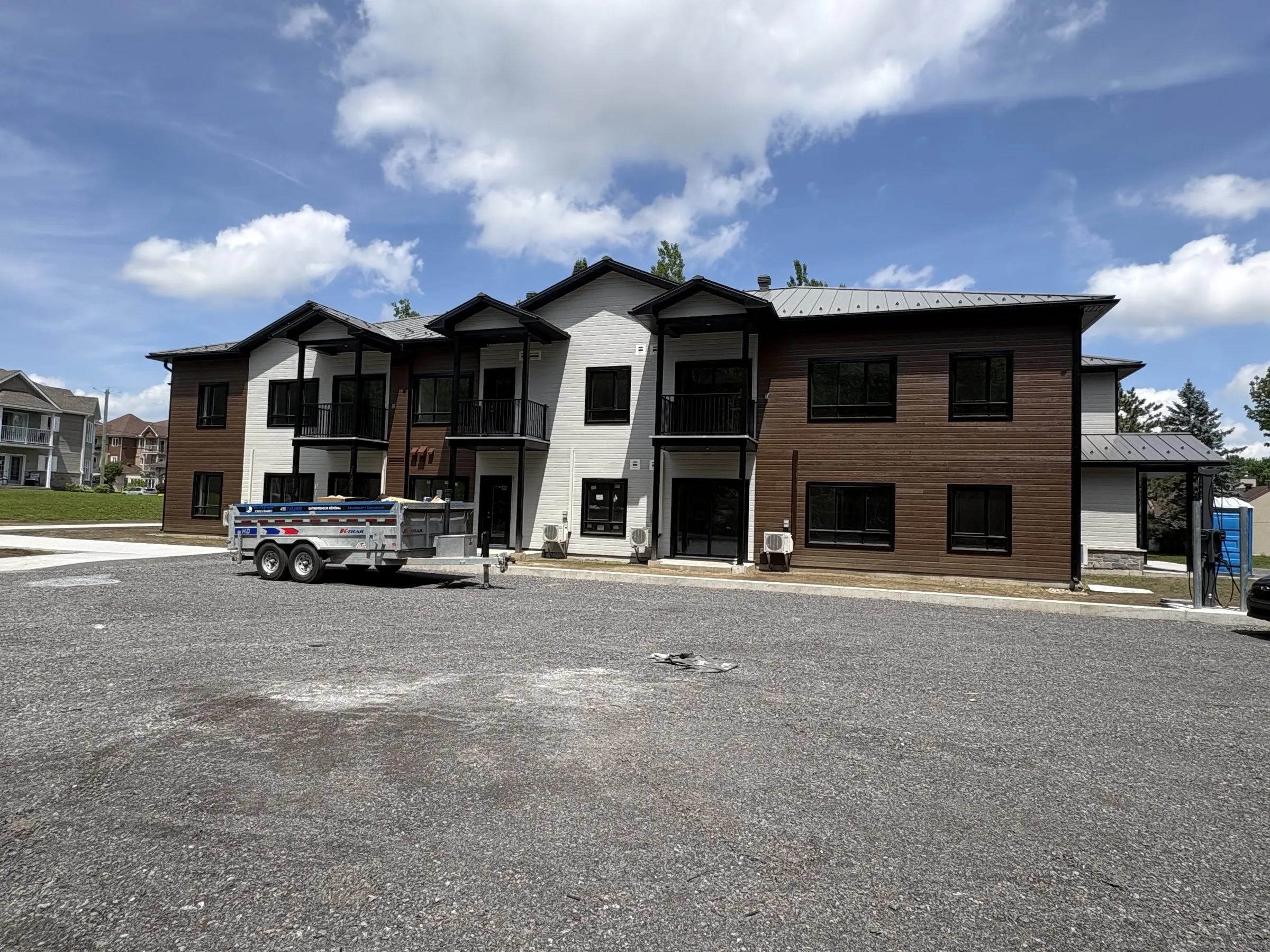 Le second immeuble en parement brun et blanc, vu du stationnement de gravier, avec les maisons du voisinage à gauche