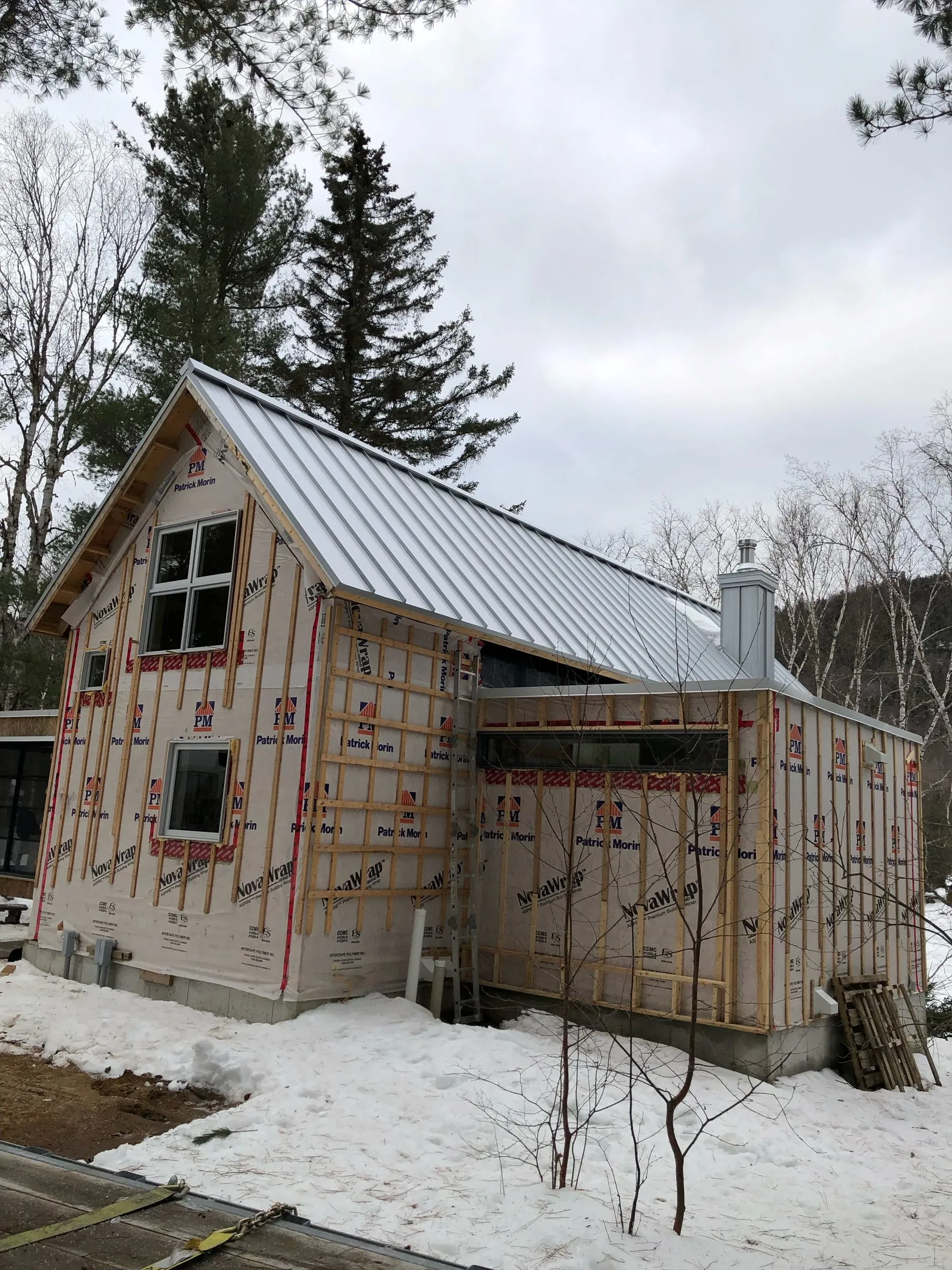 Le chalet en phase d'enveloppe, pignon et toiture métallique, vu du terrain boisé enneigé