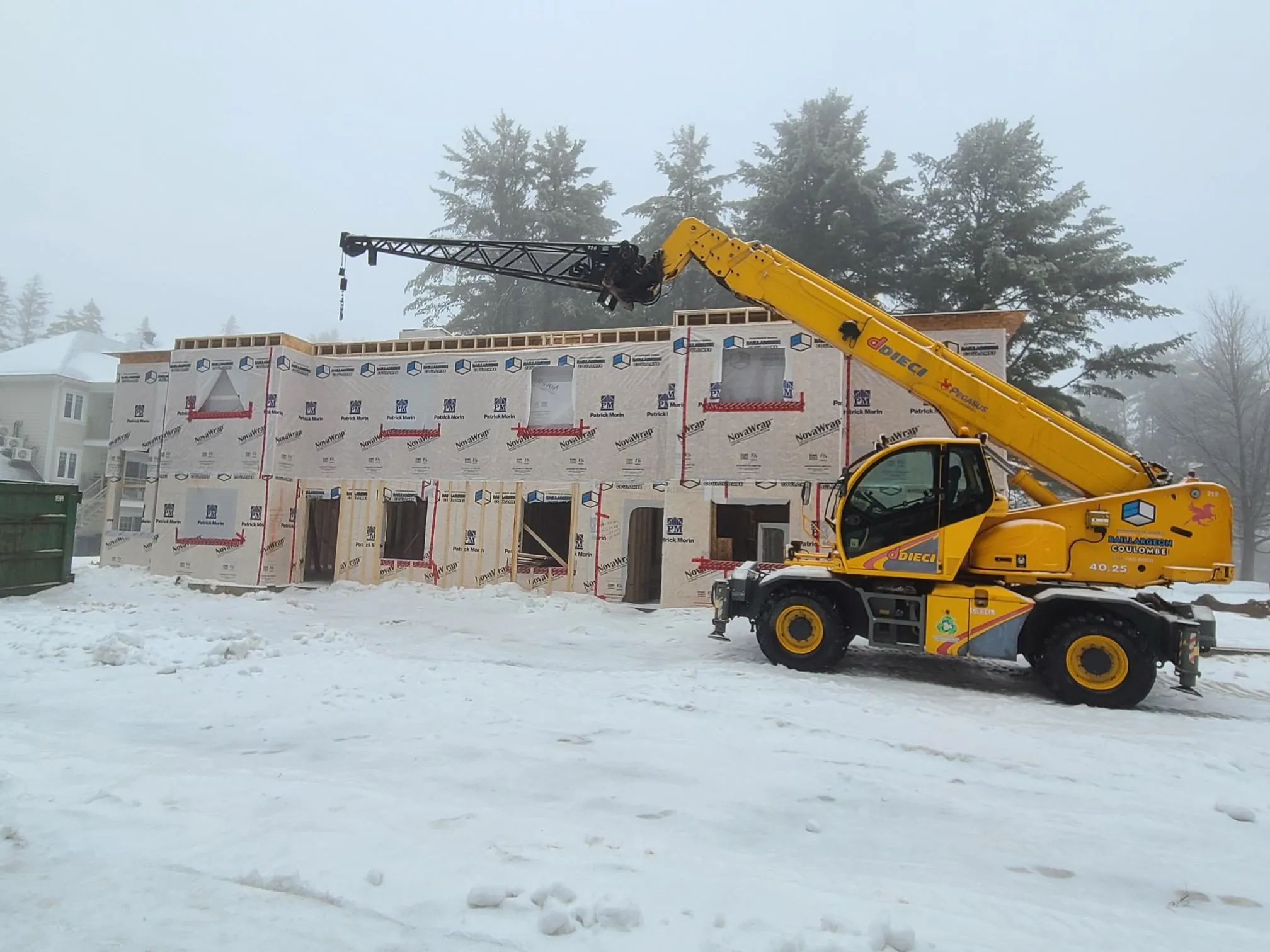 Vue large du chantier sous la neige : le bâtiment enveloppé de membrane pare-air et le chariot télescopique, flèche relevée