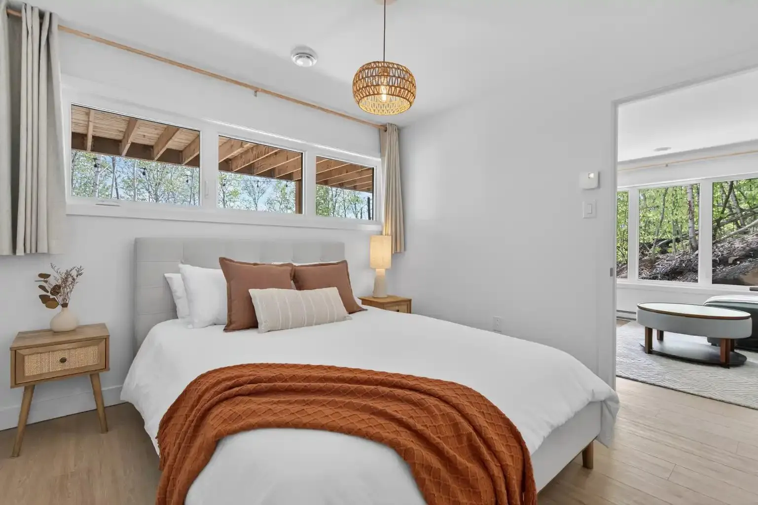 Chambre avec grand lit, table de chevet en bois et fenêtres hautes sur la forêt
