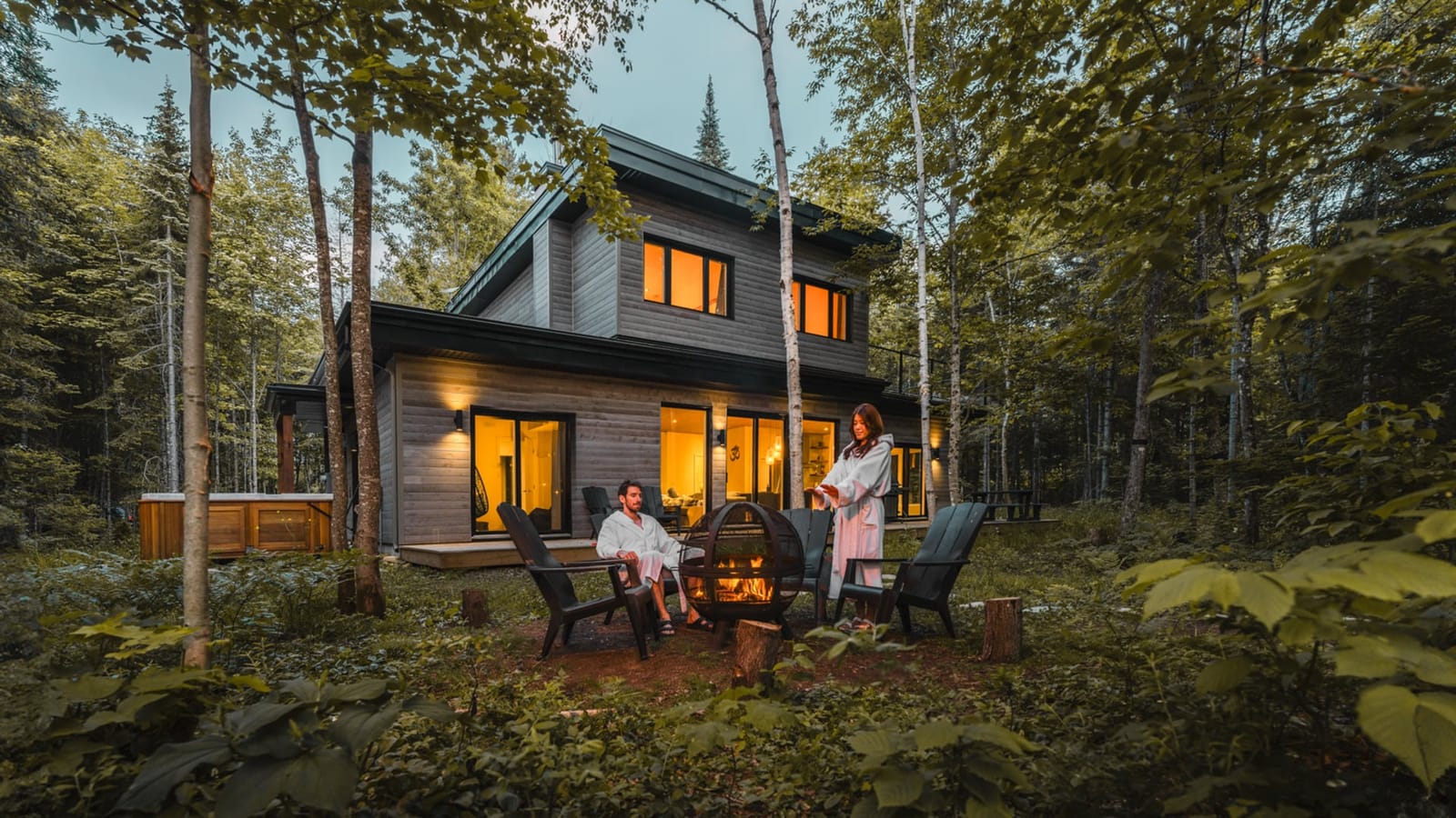 Chalet locatif haut de gamme en bois au crépuscule, avec foyer extérieur et spa nordique en forêt